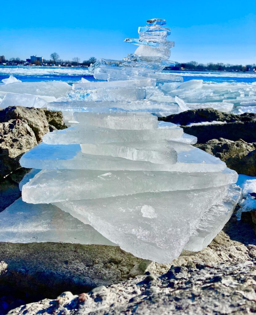On a beach, triangular pieces of ice are stacked atop each other, largest on bottom, smallest on top, wtih a sunny and ice filled Lake Michigan in the background. 